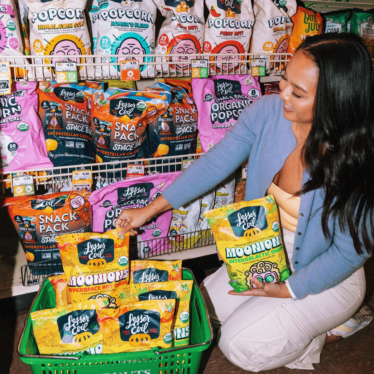 A woman kneels in front of a grocery shelf, placing bags of LesserEvil Moonion popcorn into a green Sprouts shopping basket. Other snack bags are displayed on the shelves behind her.