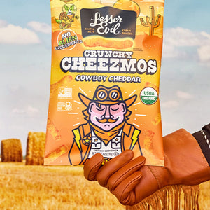 A gloved hand holds Maurices Must-haves Box by LesserEvil Snacks, with hay bales and a blue sky—perfect to enjoy alongside your finger-cleaning kit.