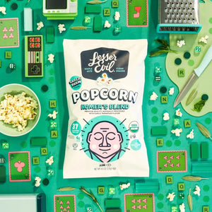 A box of LesserEvil Snacks Big Flavor Popcorn sits amid popcorn, green game boards, dice, playing cards, herbs, and kitchen utensils on a teal background.