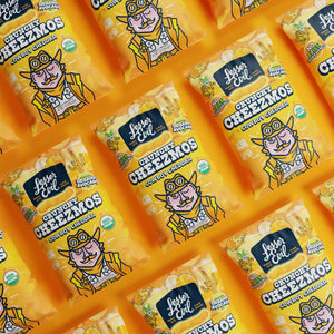 Multiple bags of LesserEvil Cowboy Cheddar Cheezmos snacks are arranged diagonally on a bold yellow background, delivering out-of-this-world flavor to your snacking mission.