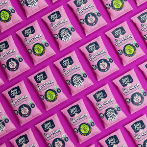 LesserEvil Snacks’ 24 Pack Snack Size Halloween Himalayan Pink Organic Popcorn bags with cartoon faces are neatly arranged on a bright pink background—ideal for Halloween parties.