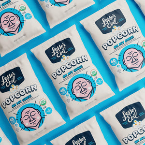 Bags of OrganicPopcorn Oh My Ghee! Gourmet Popcorn sit on a bright blue surface, showcasing their ghee flavor and fun packaging.