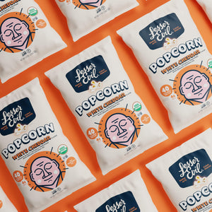 OrganicPopcorn Organic White Cheddar Flavor Popcorn bags with a cartoon face and health-focused design are arranged diagonally on an orange background.