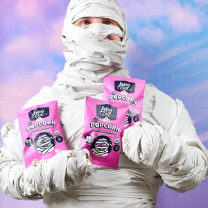 A person in a mummy costume holds LesserEvil Snacks 24 Pack Snack Size Halloween Himalayan Pink Organic Popcorn bags against a pastel sky background.