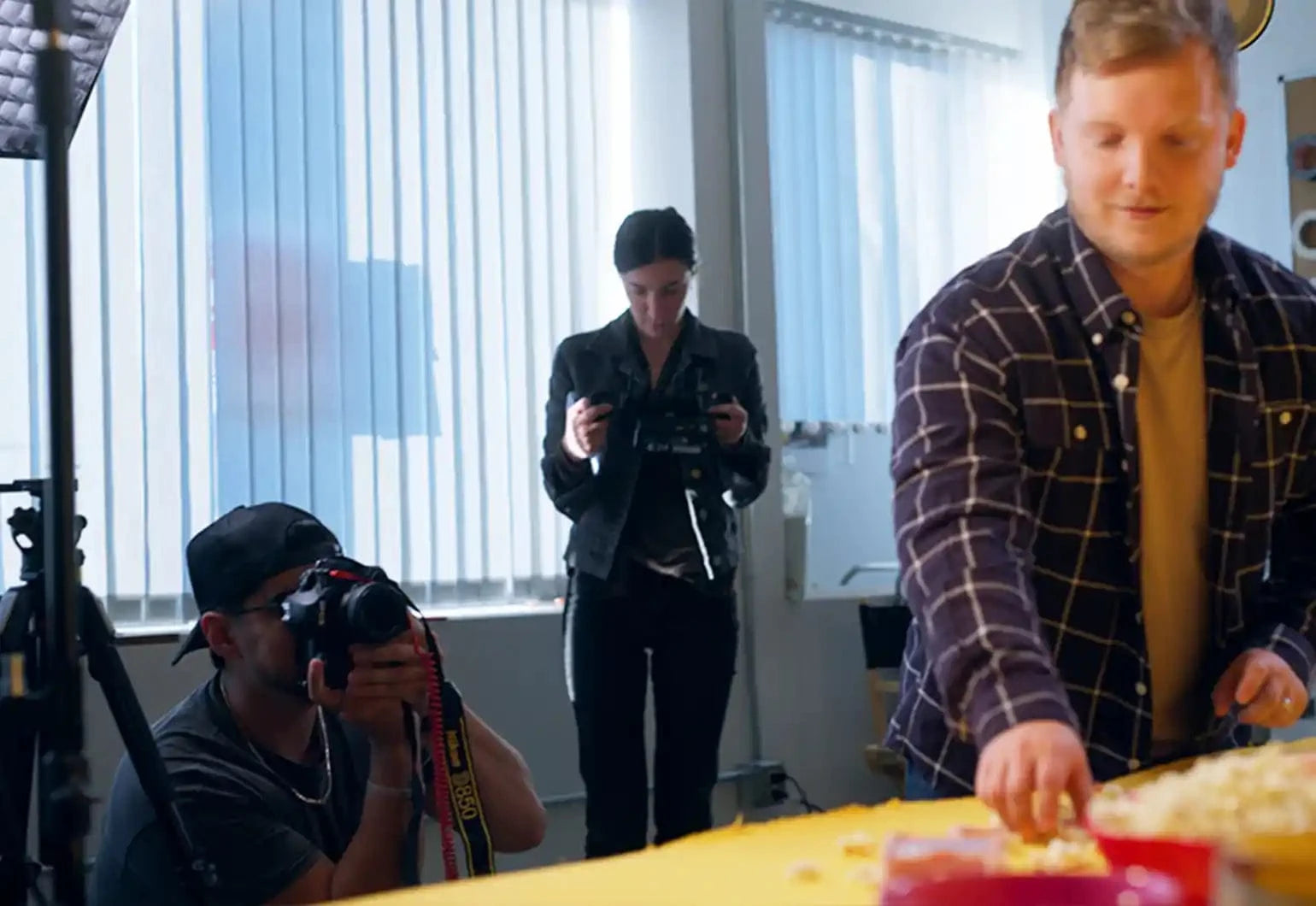 A person arranges items on a yellow table while another kneels, taking photos, and a third stands in the background looking at a camera.
