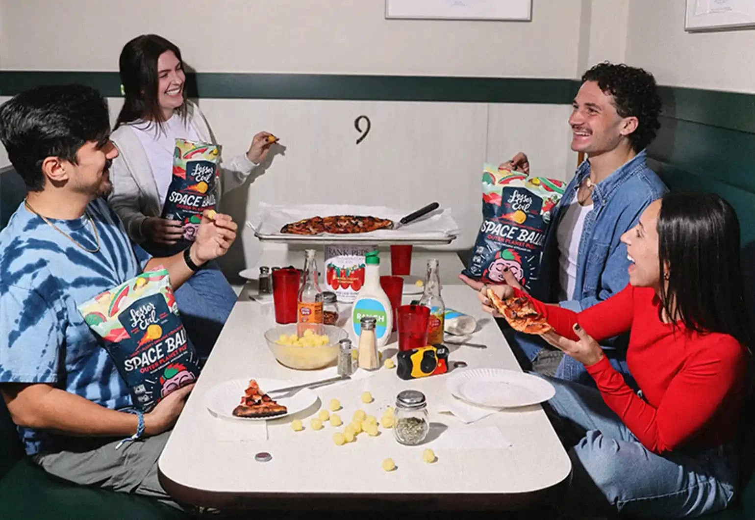 Four friends sit around a table eating pizza and snacks, smiling and laughing together. Bags of Space Balls snacks are visible on the table.