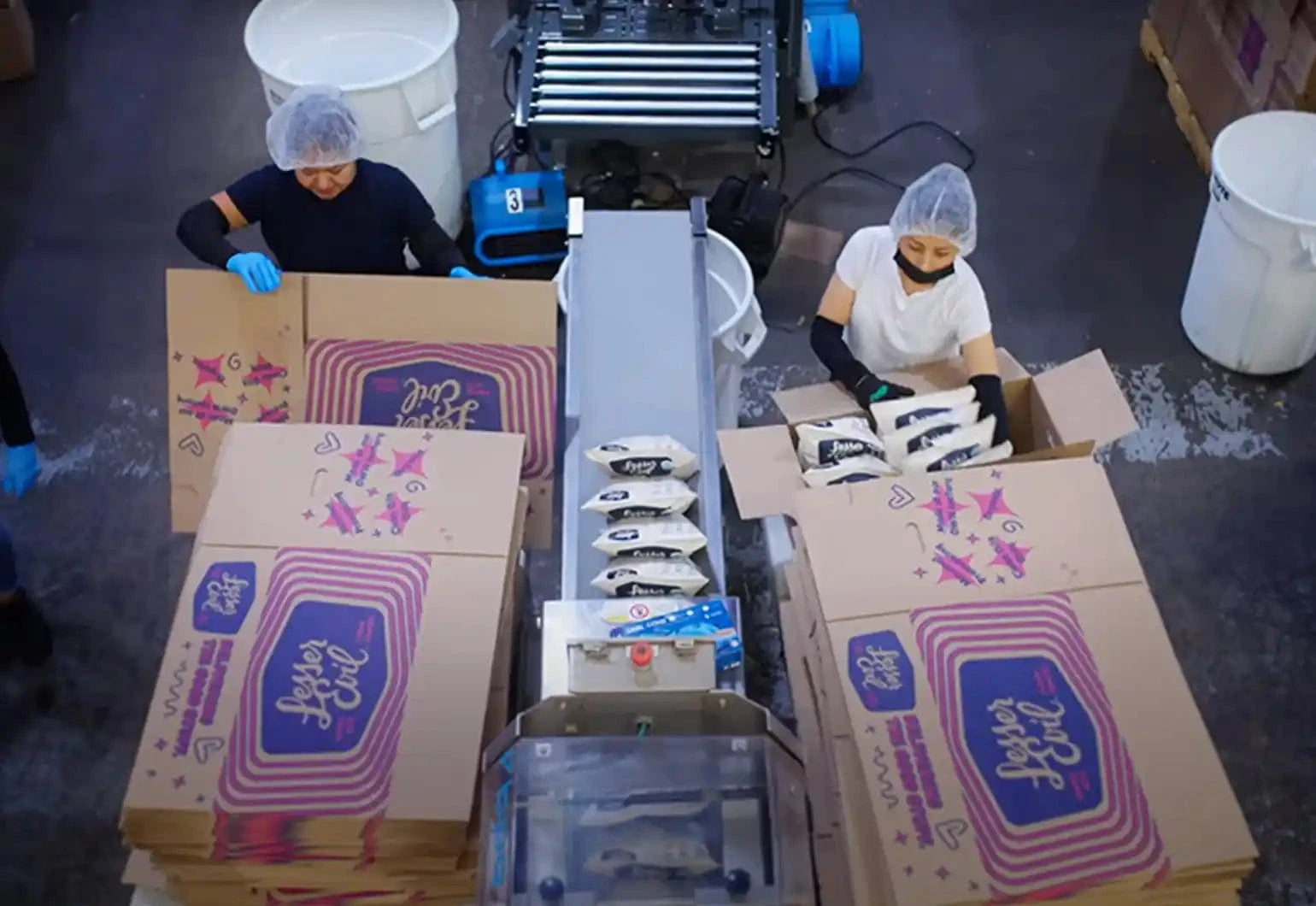 Two workers in hairnets and gloves pack food pouches into large cardboard boxes on an industrial assembly line in a factory setting.