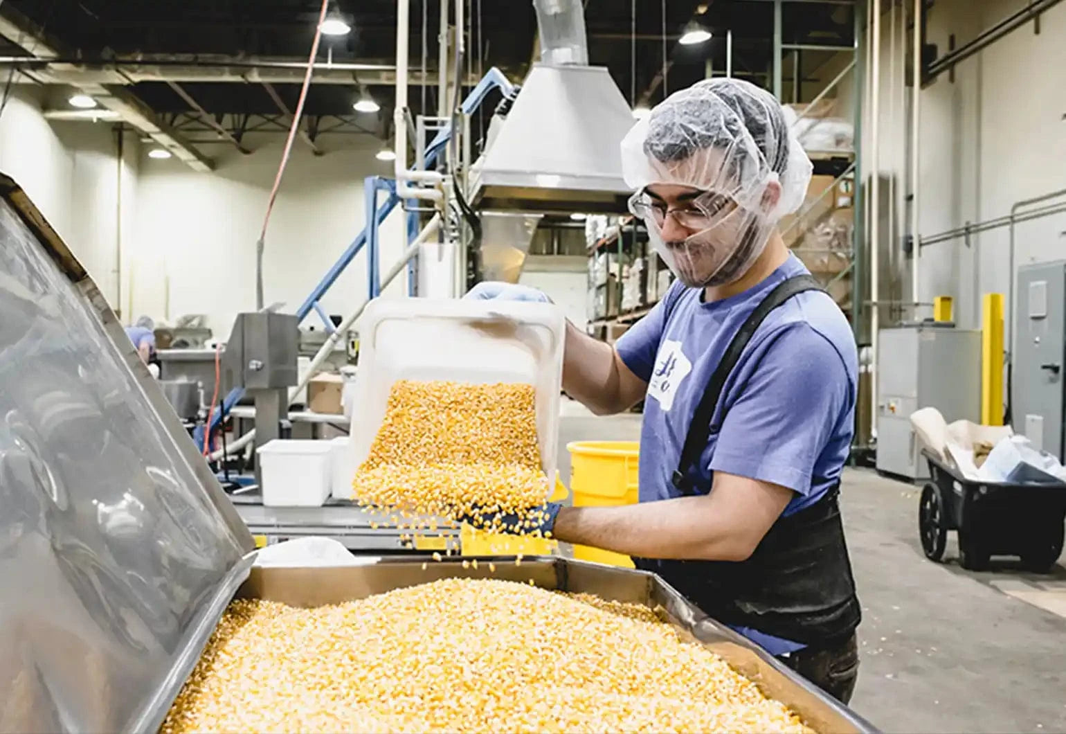 A worker in protective gear pours corn kernels into a large metal bin inside a food processing facility.