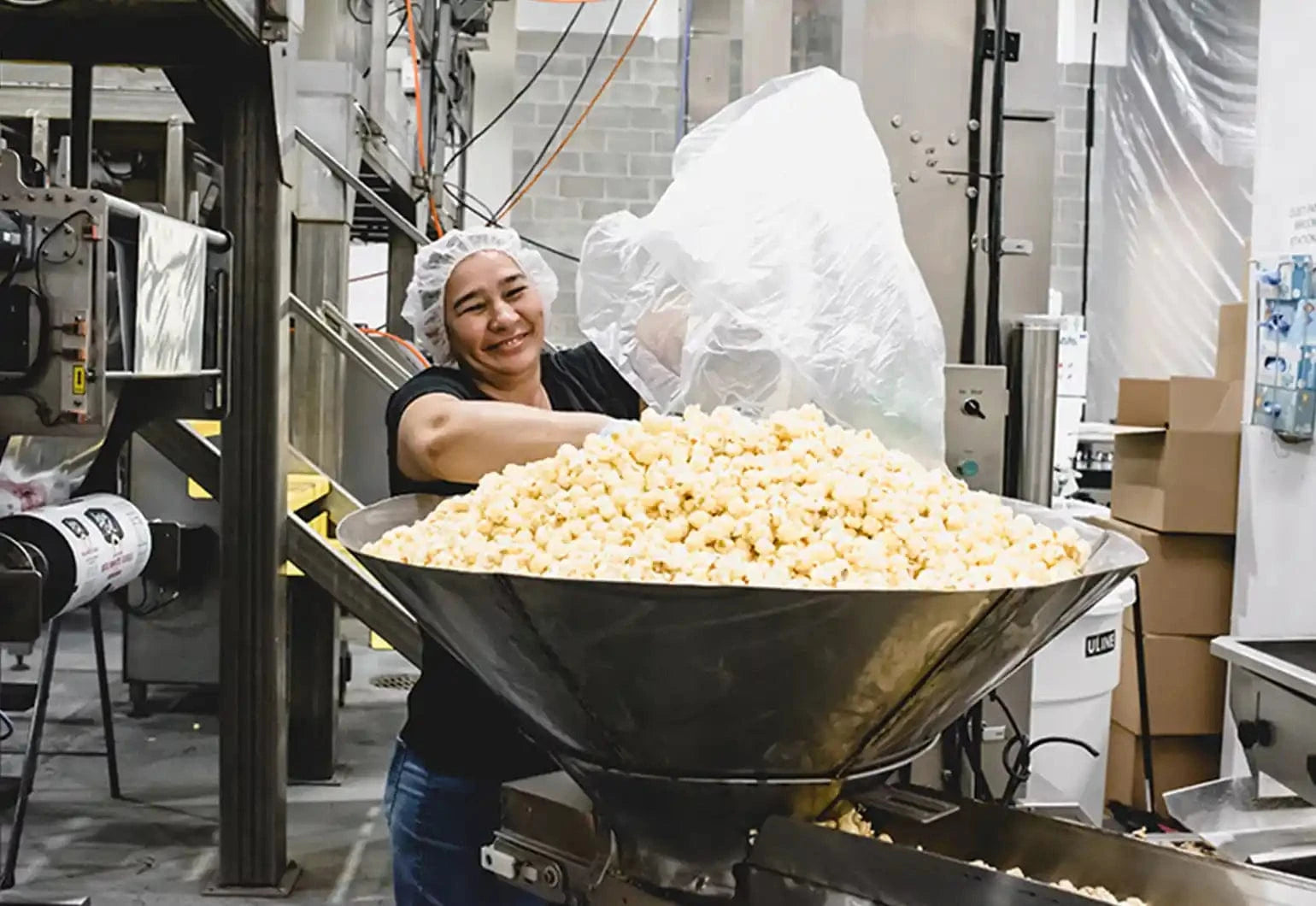 A person in a hairnet and black shirt smiles while dumping a large bag of popcorn into a metal industrial machine inside a food processing facility.
