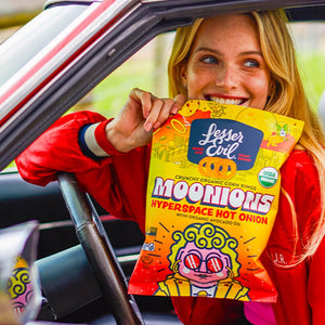 A smiling woman in a red jacket sits in a car, holding a bright yellow bag of LesserEvil Hyperspace Hot Onion Moonions made with zesty spices and avocado oil.