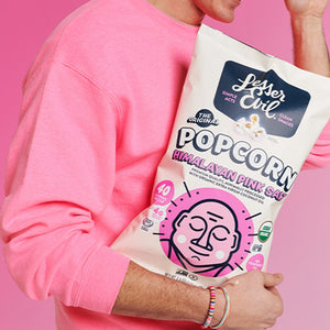 A smiling person in a pink sweatshirt holds a bag of OrganicPopcorn’s Himalayan Pink Salt Organic Popcorn against a bright pink background.