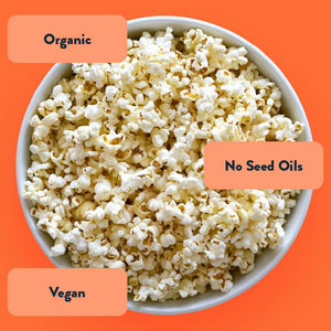 A bowl of OrganicPopcorn Organic White Cheddar Flavor Popcorn sits on an orange background, labeled organic, no seed oils, and topped with non-dairy seasoning.