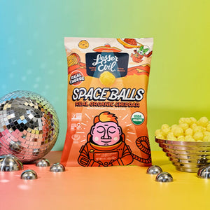 A Dairy Good Snacks Box from LesserEvil Snacks sits between a disco ball and a bowl of snacks on a colorful gradient background.