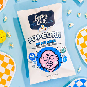 A bag of OrganicPopcorn Oh My Ghee! Gourmet Popcorn sits among pickleball paddles, balls, and popcorn on a light blue background.