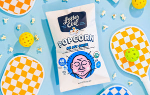 A bag of OrganicPopcorn Oh My Ghee! Gourmet Popcorn sits among pickleball paddles, balls, and popcorn on a light blue background.