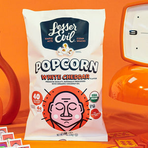 A bag of OrganicPopcorn Organic White Cheddar Flavor Popcorn sits among orange retro decor, such as clocks and vases, against a vibrant orange background.