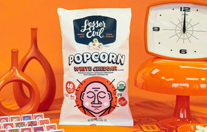 A bag of OrganicPopcorn Organic White Cheddar Flavor Popcorn sits among orange retro decor, such as clocks and vases, against a vibrant orange background.