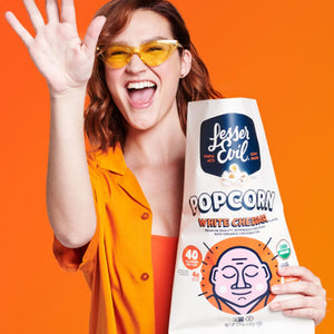 A smiling woman in yellow sunglasses and an orange shirt tosses OrganicPopcorn Organic White Cheddar Flavor Popcorn against an orange background.