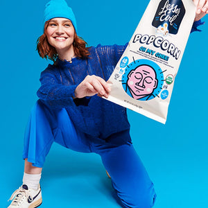 A smiling person in blue pants, sweater, and beanie poses on one knee, holding a large bag of OrganicPopcorn Oh My Ghee! Gourmet Popcorn against a bright blue background.