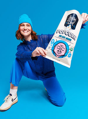 A smiling person in blue pants, sweater, and beanie poses on one knee, holding a large bag of OrganicPopcorn Oh My Ghee! Gourmet Popcorn against a bright blue background.