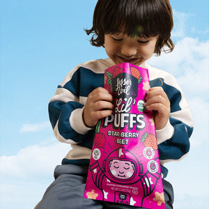 A young child smiles holding a bright pink bag of LesserEvil Star-Berry Beet Lil Puffs, a berrylicious treat with 0g added sugar, against a blue sky backdrop.