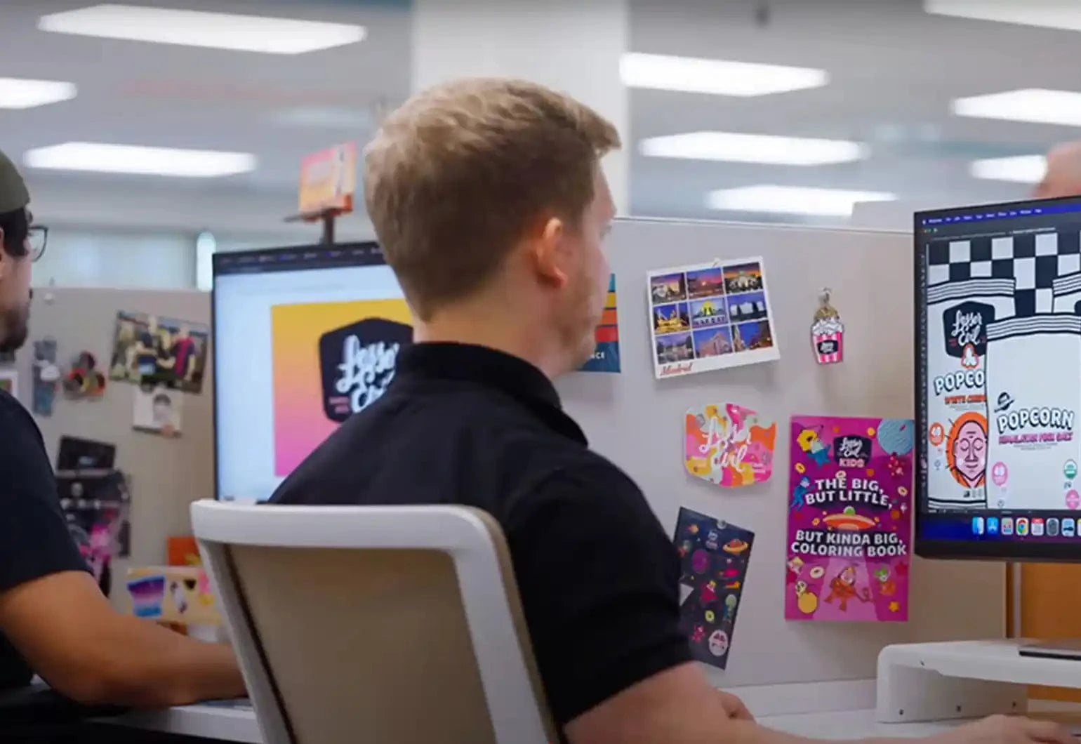 A person sits at a desk in an office, working on a computer with colorful stickers, postcards, and art pinned to the cubicle wall.