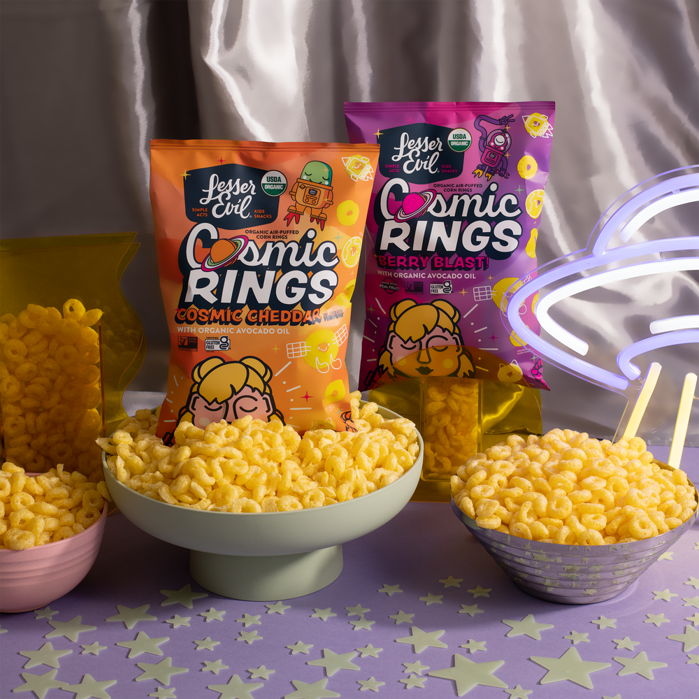 Two bags of Lesser Evil Cosmic Rings snacks (Cosmic Cheddar and Berry Blast) stand behind bowls filled with the ring-shaped snacks on a purple table with stars and a glowing neon cloud sign.