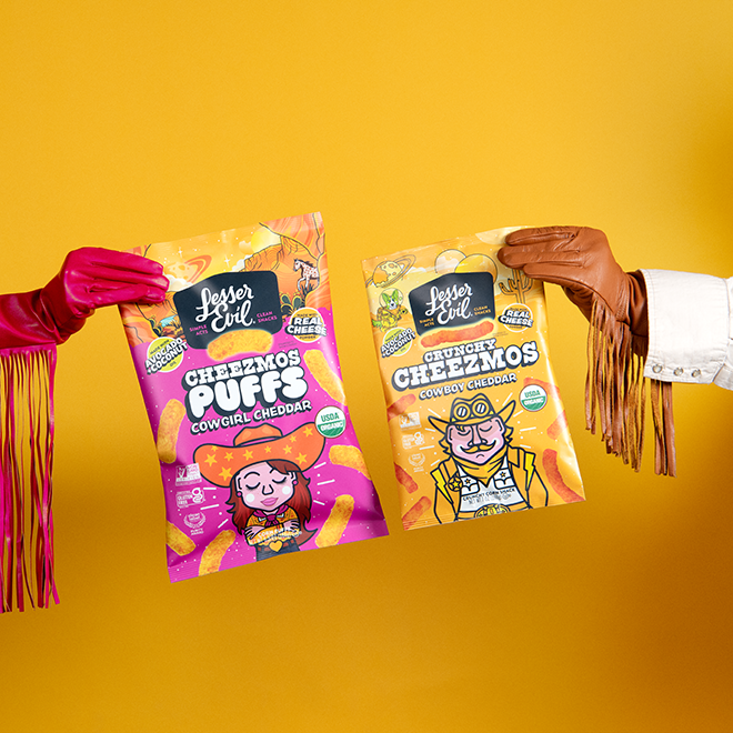 A person in a red embroidered shirt holds a bag of Lesser Evil Crunchy Cheezmos Blazin Hot Cheddar snack, featuring a cartoon aviator cat and southwestern-themed packaging.