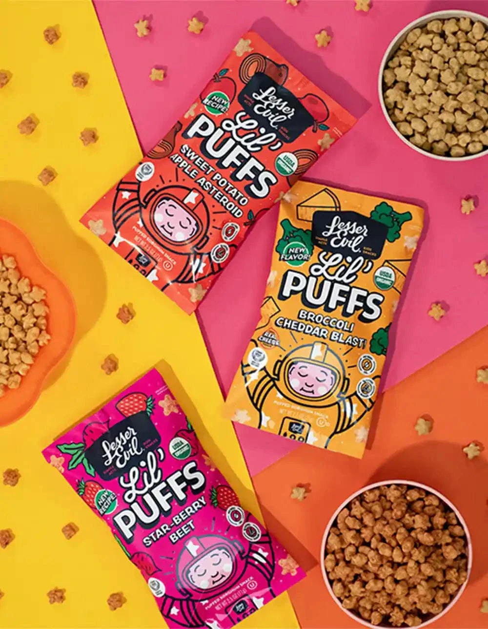 Three colorful bags of Lesser Evil Lil Puffs snacks in Sweet Potato, Broccoli Cheddar Blast, and Star-Berry flavors are on a pink background with small bowls and scattered puff snacks.