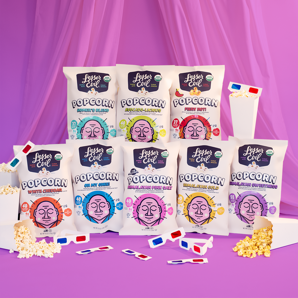 Eight bags of LesserEvil popcorn in various flavors are arranged on a pastel purple background with scattered popcorn, popcorn boxes, and 3D glasses in the foreground.