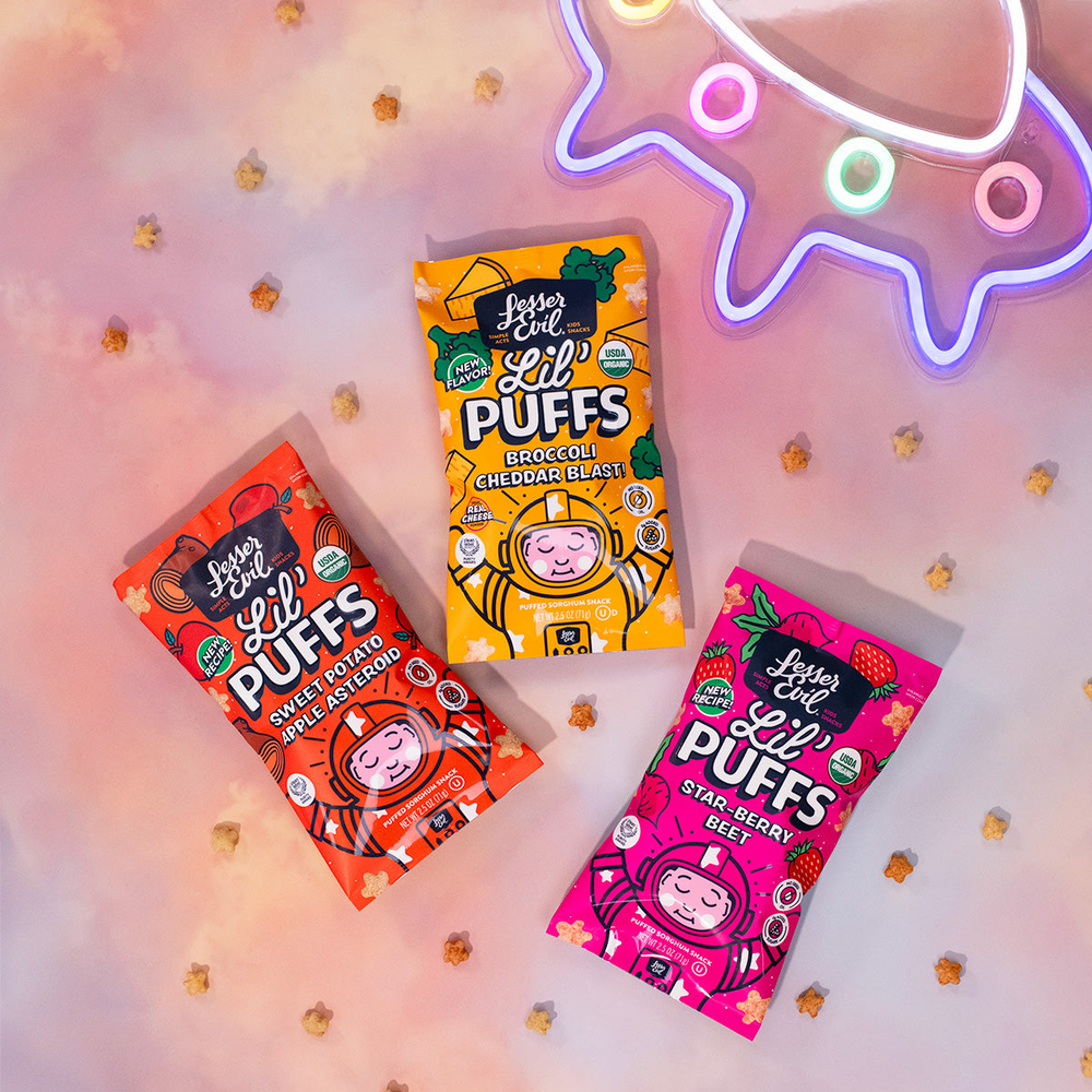Three colorful bags of Lesser Evil Lil Puffs snacks (Sweet Potato Apple, Broccoli Cheddar Blast, Star-Berry Beet) are arranged on a pastel background with scattered puffs and a neon UFO sign.