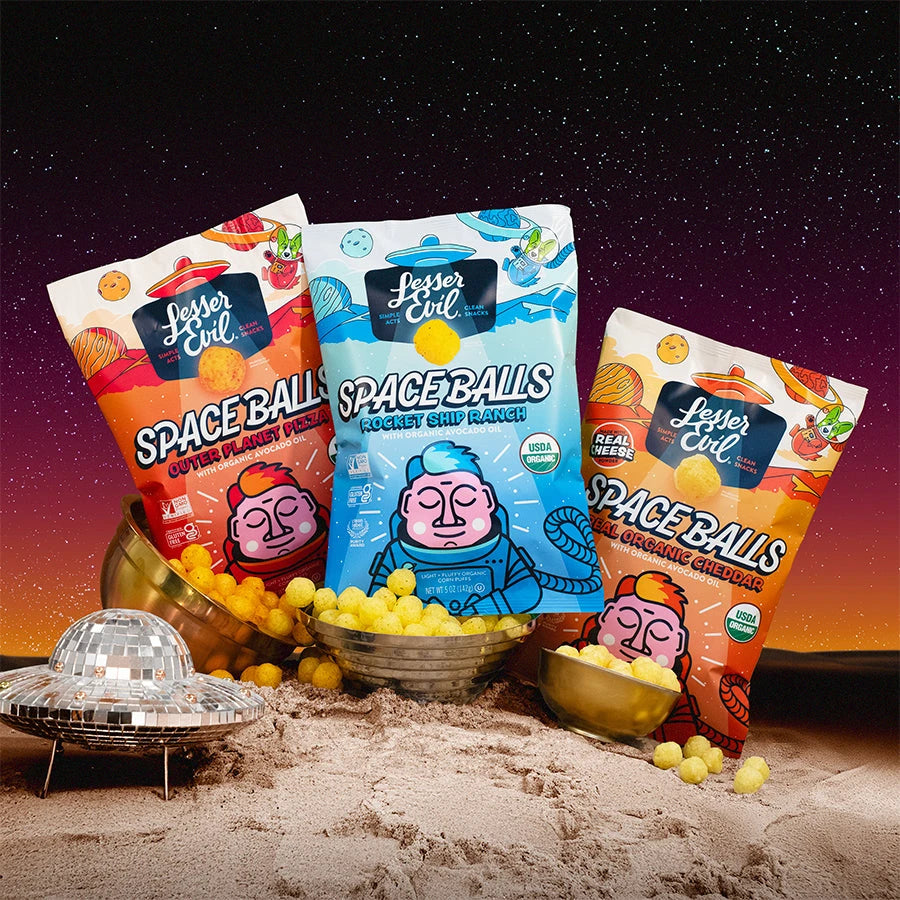 Three colorful bags of LesserEvil Space Balls snacks sit on sandy terrain with cheese balls spilling out, a small disco-ball UFO nearby, and a vibrant space-themed background.