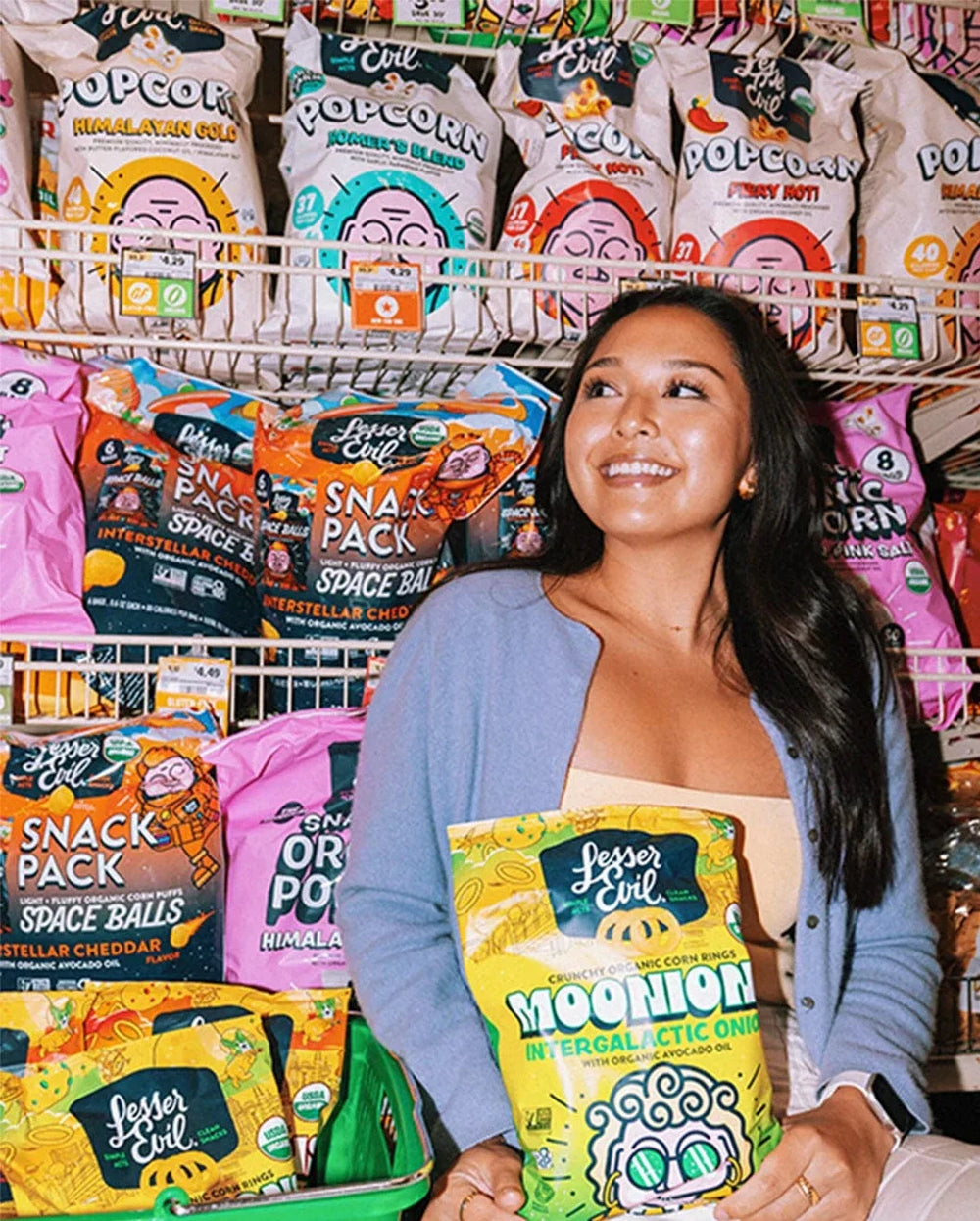 A woman smiles while holding a bag of Lesser Evil Moonion snacks, standing in front of shelves filled with colorful popcorn and snack bags.