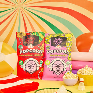 Two brightly colored bags from the Sweet Summer Box by LesserEvil Snacks sit on a yellow table, surrounded by popcorn, rackets, and a swirl backdrop for juicy summer flavors.