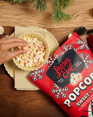 A hand reaches for Sweet + Salty Popcorn in a bowl beside a red LesserEvil bag—an ideal holiday snack—on a festive, pine-decorated wooden table.