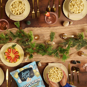 A festive table with gold cutlery, pink plates, strawberries, and LesserEvil Snacks’ Holiday Popcorn Box in sweet & salty white chocolate marshmallow. Blue sleeves hold a bowl below.
