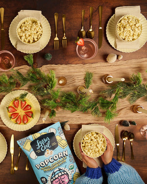 A festive table with gold cutlery, pink plates, strawberries, and LesserEvil Snacks’ Holiday Popcorn Box in sweet & salty white chocolate marshmallow. Blue sleeves hold a bowl below.