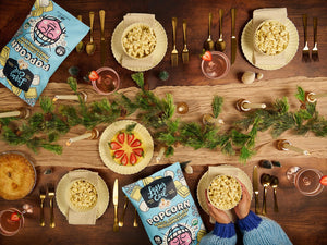 A festive table with LesserEvil White Chocolate Marshmallow popcorn, pie, drinks, and pine garland—golden cutlery adds charm to this perfect holiday snack scene in cozy blue sweaters.