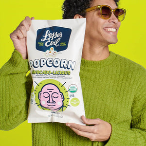 A smiling person in sunglasses and a green sweater holds a bag of LesserEvil Avocado-licious Organic Popcorn against a bright yellow background.