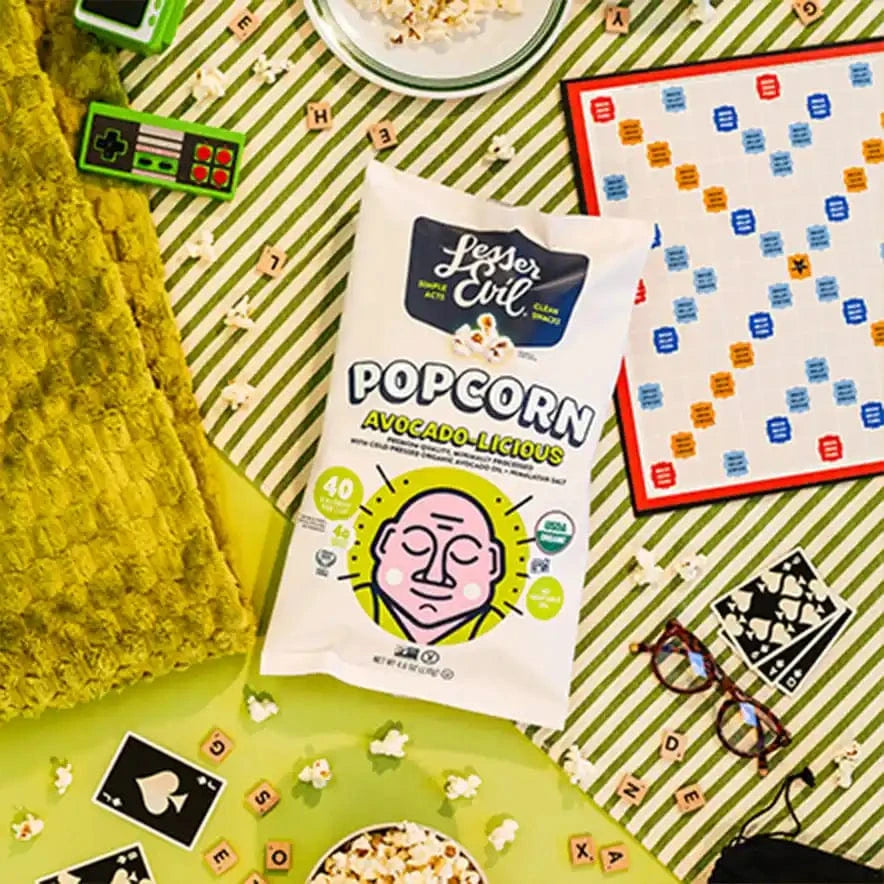 A bag of The Classics Popcorn Box by LesserEvil Snacks sits on a striped green table with popcorn, games, cards, sunglasses, and a yellow textured blanket.