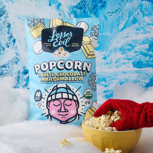 A gloved hand holds sweet popcorn in a yellow bowl beside a LesserEvil White Chocolate Marshmallow bag on fake snow with snowy mountains in the background.