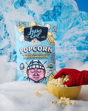 A gloved hand holds sweet popcorn in a yellow bowl beside a LesserEvil White Chocolate Marshmallow bag on fake snow with snowy mountains in the background.