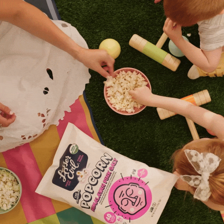 Three children and an adult sitting on grass share a bowl of popcorn during a picnic, with a colorful blanket, croquet set, and a bag of popcorn visible.