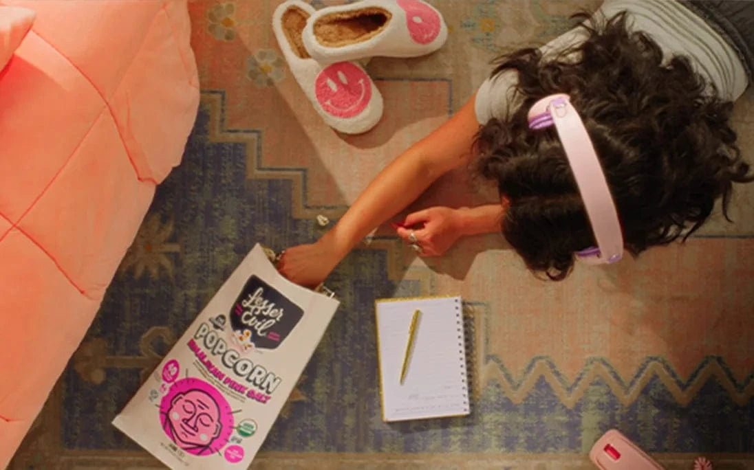 A person wearing headphones lies on a rug, reaching for popcorn. Nearby are slippers, a notepad with a pencil, and a popcorn bag with playful designs.