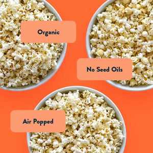 Three bowls of popcorn on orange background labeled Organic, White Cheddar, and Air Popped from The Classics Popcorn Box by LesserEvil Snacks.