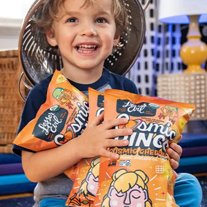 A smiling child wears a metal colander as a hat and holds two bags of LesserEvil Cosmic Cheddar Cosmic Rings vegan snacks while sitting indoors.
