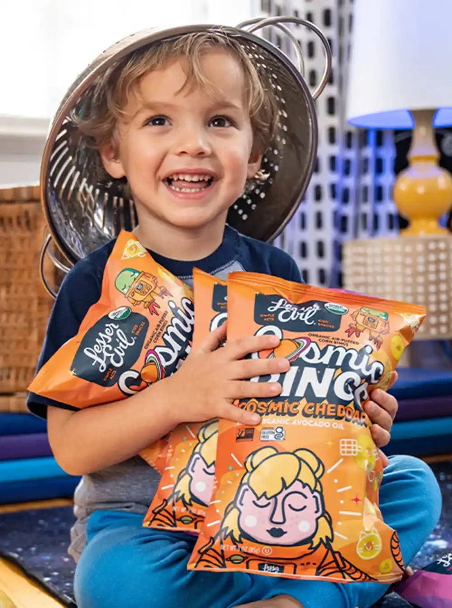 A smiling child wears a metal colander as a hat and holds two bags of LesserEvil Cosmic Cheddar Cosmic Rings vegan snacks while sitting indoors.