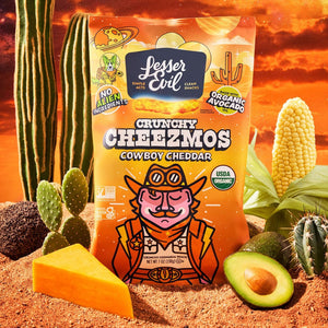 A Cheesy Snack Box from LesserEvil Snacks sits in a desert with cacti, corn, cheese, and avocado under an orange sky. Try it today!.