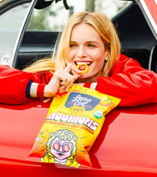 A smiling woman in a red jacket leans out of a car window, eating a chip from a yellow bag labeled Moonions Hyperspace Hot Onion.