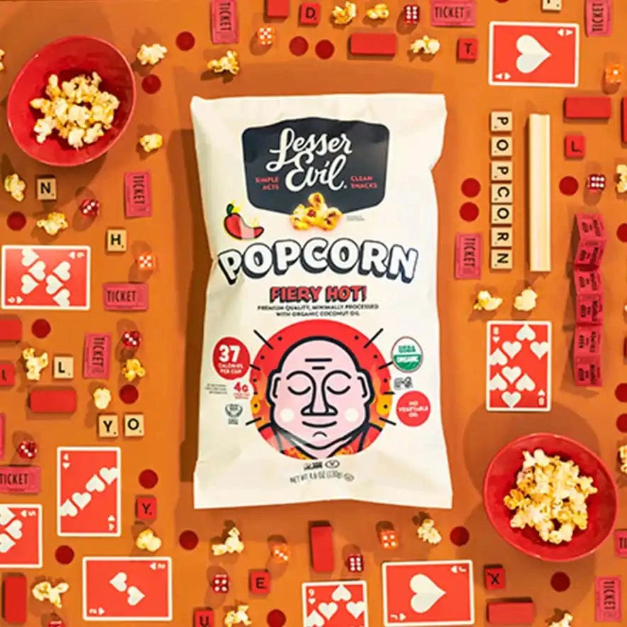 A Little Spice Snacks Box by LesserEvil Snacks rests on an orange surface with bowls, cards, tickets, dice, and popcorn—an ideal game night treat.